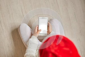 Top view of little child with smartphone with blank screen in hands hands, kid wearing santa hat, white pullover and tights, Copy