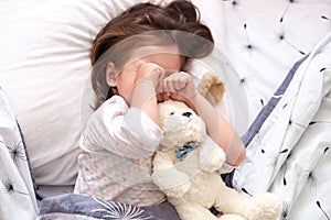 Top view of little girl lying in bed with teddy bear, being in bad mood, does not want to srtand up and go to kinder garten,
