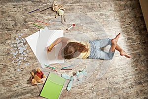 Top view of little girl lying on the wooden floor in her room at home and drawing with colorful pencils on a white sheet