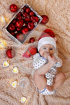 Top view on a little girl in Santa Claus red hat is laying on a beige plaid with red and white Christmas decorations.