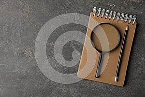 Top view of magnifier glass, notebook and pencil on grey stone background, space for text. Find keywords concept