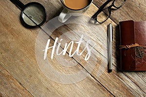 Top view of magnifying glass, coffee, glasses, pen and notebook on wooden background written with Hints