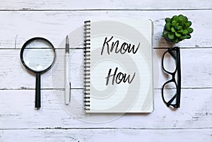 Top view of magnifying glass,glasses,plant,pen and notebook written with Know How on white wooden background.