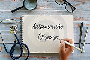 Top view of magnifying glass,glasses,stethoscope,pen and hand writing ` Autoimmune Disease ` on notebook on wooden background photo