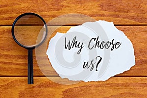 Top view of magnifying glass and piece of paper written with question Why Choose Us? on wooden background.