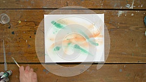 Top view of male artist paints an abstract picture, he uses green paint on wet canves and distribute it with paintbrush