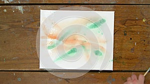 Top view of male artist paints an abstract picture, he uses green paint on wet canves and distribute it with paintbrush