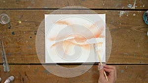 Top view of male artist paints an abstract picture, he uses orange paint on wet canves and distribute it with paintbrush