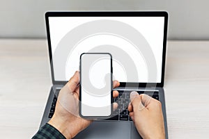 Top view of male hands holding smartphone with mockup on screen beside laptop. background of wooden table.