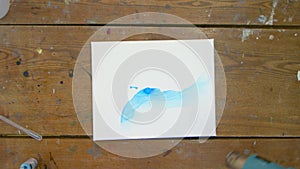 Top view of man artist paints beautiful abstract picture, he pours blue paint from tube on wet canvas uses dryer to move