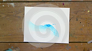 Top view of man artist paints beautiful abstract picture, he pours blue paint from tube on wet canvas uses dryer to move
