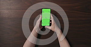 Top view man hands hold smartphone with green screen over black walnut wood table