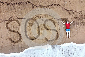 Top view on man laying on desert beach close to big inscription on the sand SOS.