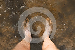 Top view of a man standing barefoot in muddy water, close-up of male bare feet in sand and mud. Muddy water, dirty sea