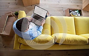 Top view of mature man sitting on sofa in unfurnished house, using laptop.