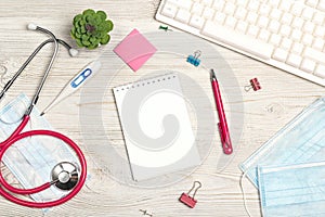 Top view medical workplace with blank notepad, stethoscope, keyboard, thermometer, mask, plant and supplies on white wooden desk