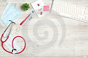 Top view medical workplace with stethoscope, keyboard, thermometer, mask, plant and supplies on white wooden desk