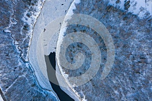 Top view of melting ice in a flowing river through the snow-covered forest on a winter day
