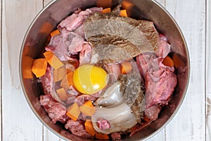 Top view of metal pet bowl with chicken necks, beef meat, paunch, raw egg and carrot pieces. Natural organic daily meal ration pre