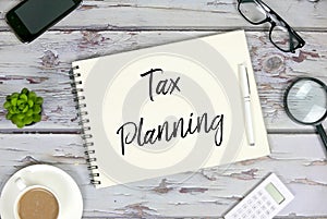 Top view of mobile phone,plant,coffee,calculator,magnifying glass,sunglasses,pen and notebook written with Tax Planning on wooden