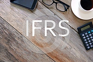 Top view of mobile phone, sunglasses, a cup of coffee and calculator on wooden background written with IFRS