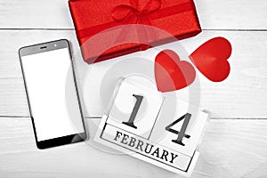 Top view of mobile smart phone with white blank screen, red gift box, hearts and wood block calendar on wood background