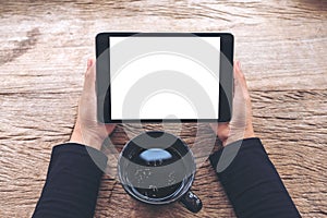 Top view mockup image of hands holding black tablet pc with blank white screen and coffee cup on vintage wooden table