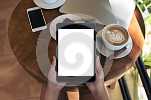 Top view mockup image of hands holding black tablet pc with blank white screen , smart phone , laptop , coffee cup and cake