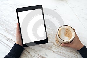 Hands holding black tablet pc with white blank screen and coffee cup on table background