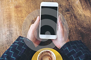 Top view mockup image of hands holding white mobile phone with blank black desktop screen and yellow coffee cup on wooden table