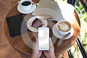 Top view mockup image of hands holding white smartphone with blank screen , tablet , laptop , coffee cup and cake