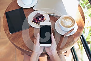 Top view mockup image of hands holding white smartphone with blank screen , tablet , laptop , coffee cup and cake on wooden table