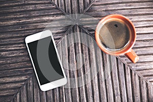 Top view mockup image of a white mobile phone with blank black desktop screen and a cup of coffee on vintage wooden table