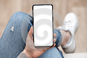 Top view mockup image of woman holding mobile phone with blank white screen. Woman in jeans sits cross-legged and holds cell phone