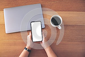 A woman holding and using black mobile phone with blank white desktop screen with coffee cup and laptop