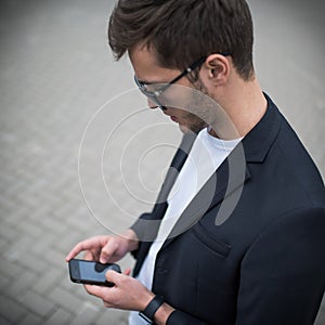 Top view of modern young man wearing black suit and sunglasses, texting on the phone messages for his girlfirend in the street.