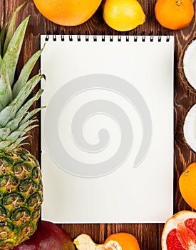 top view of note pad with pineapple orange lemon tangerine coconut mango grapefruit around on wooden background with copy space
