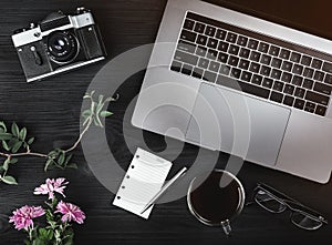 Top view of notebook computer, eyeglasses, cup of coffee and white sheet, vintage camera and pink flowers