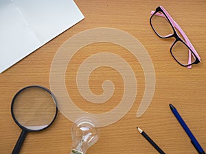 Top view of notebook, light bulb, pink glasses, Magnifying glass pen and pencil on wooden table with center copy space