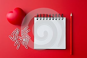 Top view of Notebook, pencil, paper clip, heart shaped ball  on a red background and copy space