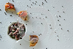 Top view of octopus salad with slices of toasted baguette - bruschetta on white background with black pepper and sea salt.