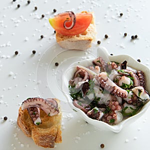 Top view of octopus salad with slices of toasted baguette - bruschetta on white background with black pepper and sea salt.