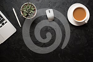 Top view of office desk. Green plant in a pot, cup of coffee, computer mouse, pen and modern silver laptop on dark background.