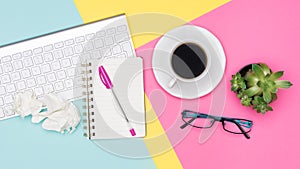 Top view office desk with notepad, wireless keyboard, succulent plant, coffee cup and glasses on pastel colored background.