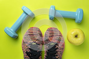 Top view of one apple , two sneakers and two dumbbells on green background.Fitness equipment with healthy food.Sport accessories