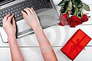 Top view of open laptop computer, bouquet of roses, gift box and woman hands typing on keyboard. Valentines day, Womans day