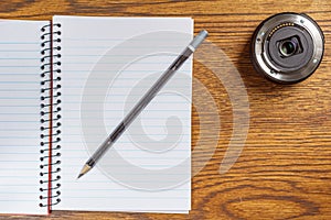 Top view of open spiral notebook with black pencil and camera lens on desk background.