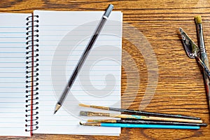 Top view of open spiral notebook with black pencil and paint brushes on desk background.