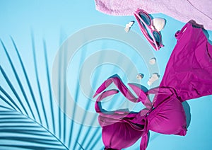 Top view of palm leaves, swimsuit, dried shells, shades, and a pink towel on a blue background