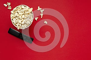 Top view of paper bucket full of sweet popcorn, black tv clicker on the red background.Empty space